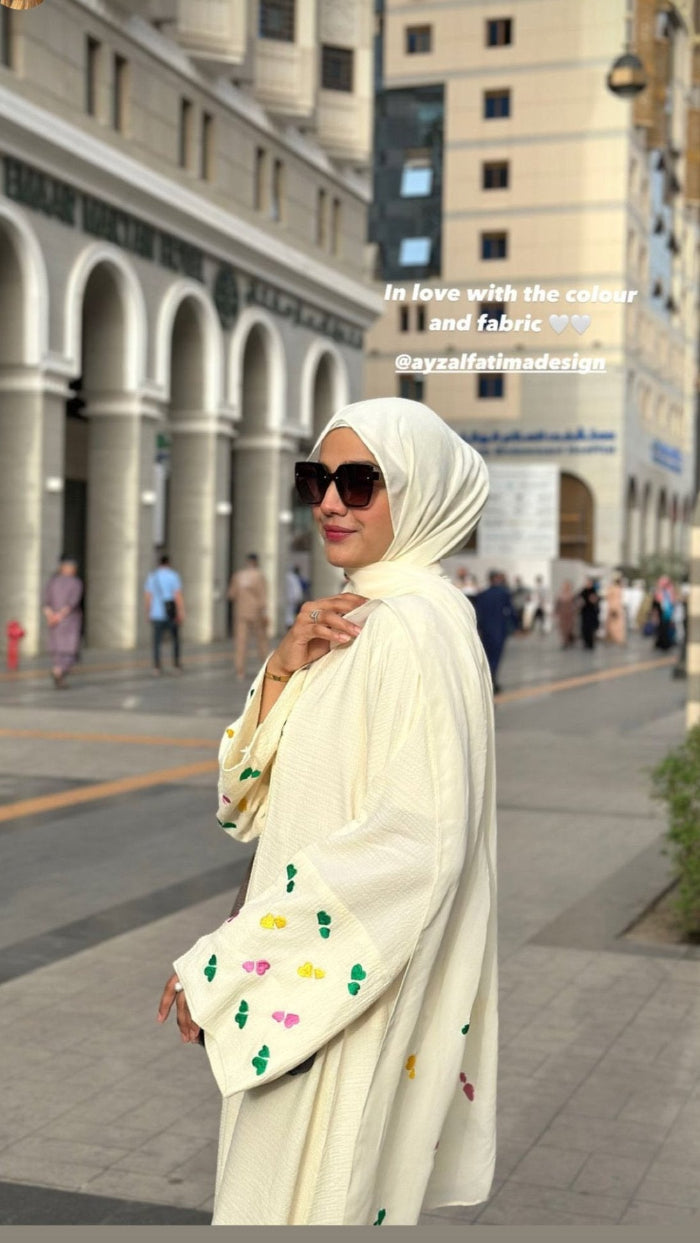 A woman wearing a white traditional abaya with a floral pattern, sunglasses, and a lemon-yellow color.