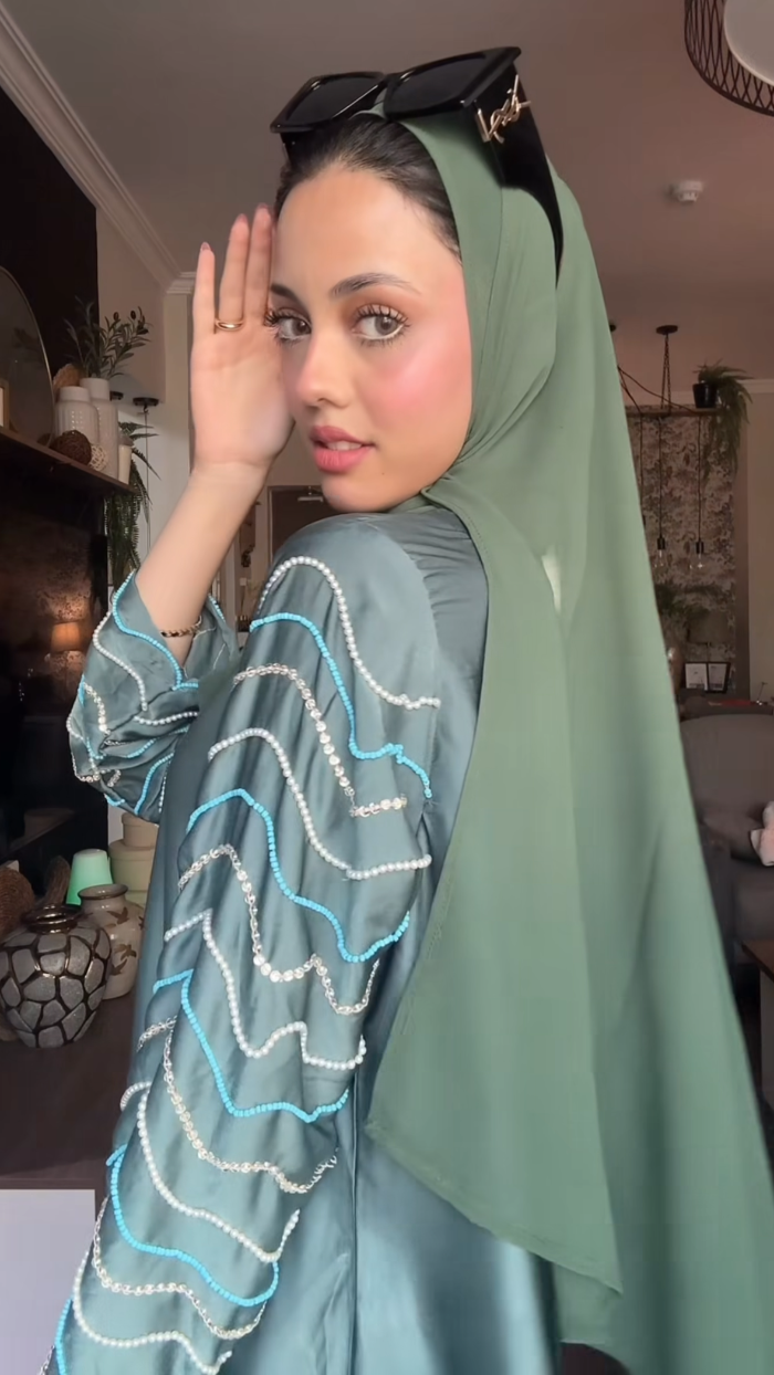 Woman wearing a green hijab and light blue dress with white patterns indoors.