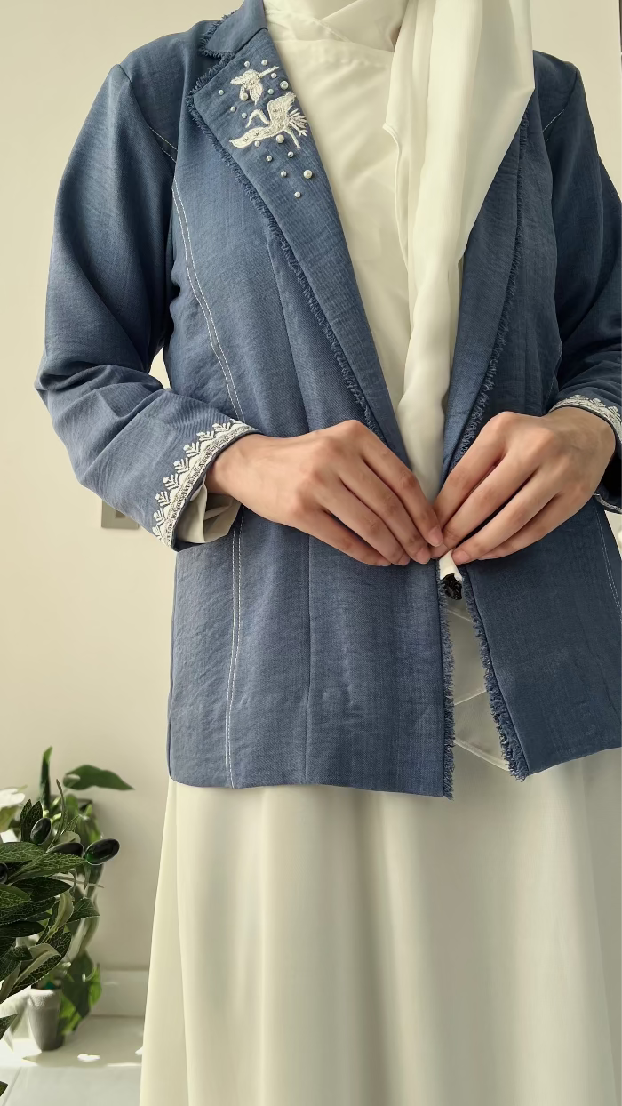 Person wearing a blue embroidered jacket with a white background and plant on the side