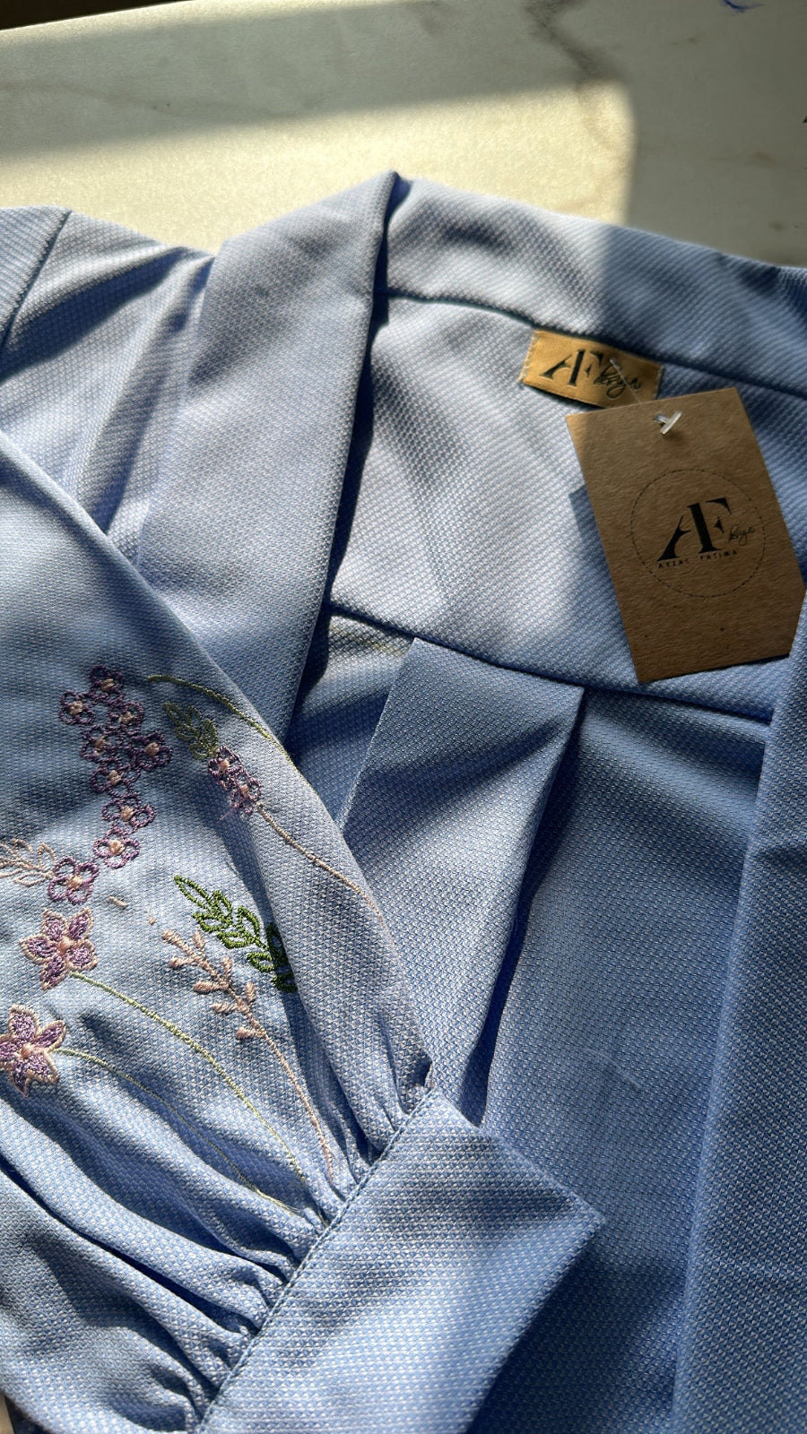 Light blue fabric with floral embroidery and a brand tag on a neutral background