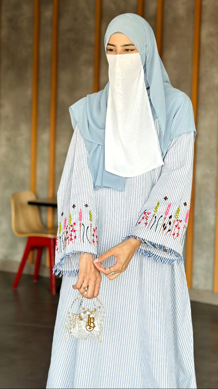 Woman wearing a light blue hijab and dress with floral embroidery in an indoor setting.