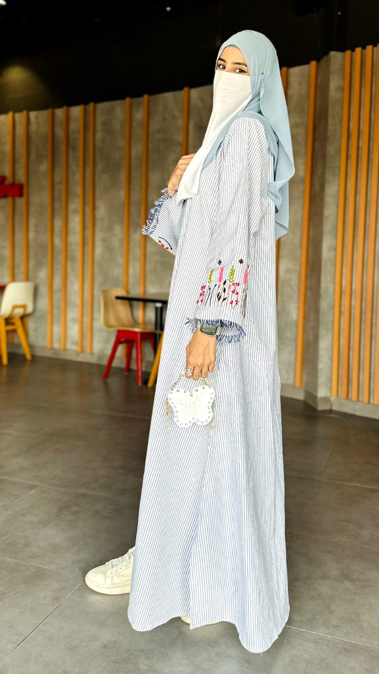 Woman in a long light gray dress with floral embroidery standing in a modern indoor setting.