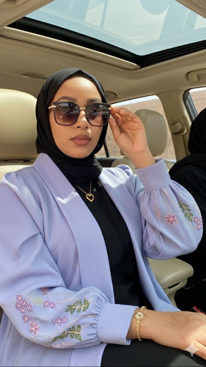 Woman sitting in a car wearing a light blue embroidered cardigan and sunglasses.