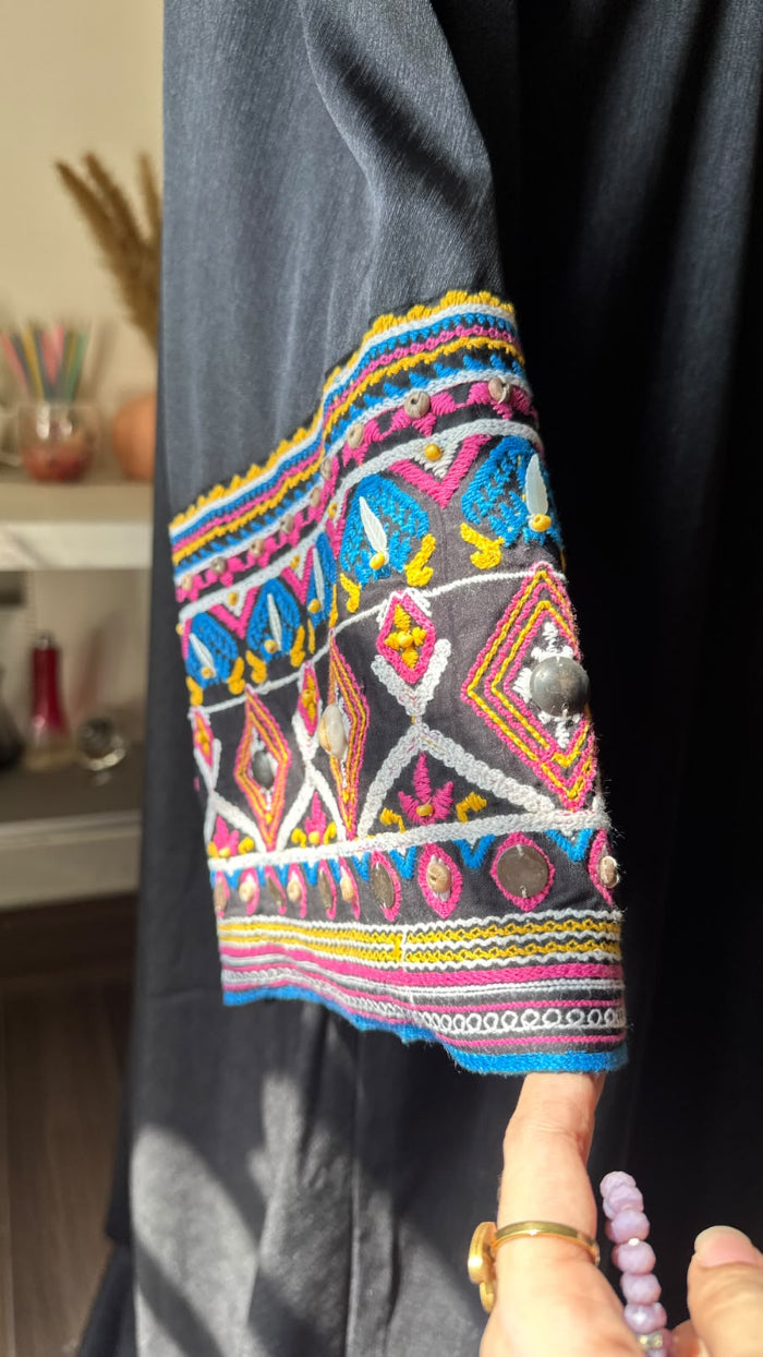 A close-up of a dark charcoal gray garment featuring a highly colorful, geometric embroidered sleeve cuff with pink, blue, yellow, and white patterns, held by a hand wearing a beaded bracelet.