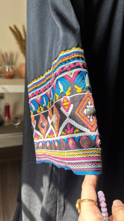 A close-up of a dark charcoal gray garment featuring a highly colorful, geometric embroidered sleeve cuff with pink, blue, yellow, and white patterns, held by a hand wearing a beaded bracelet.