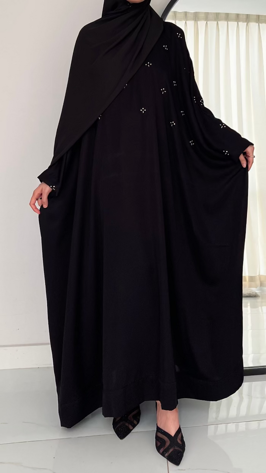 Person wearing a black abaya with decorative buttons in a minimal indoor setting.
