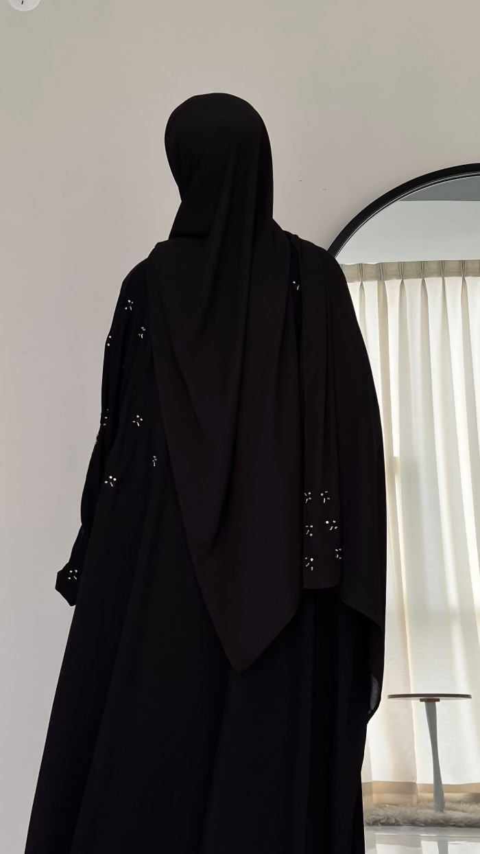 Person wearing a black abaya standing in front of a mirror with a light curtain.