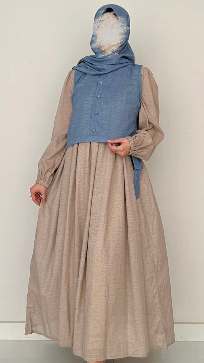 Person wearing a long beige dress with a blue denim vest and headscarf on a plain background