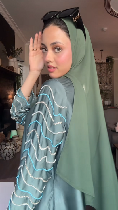 Woman wearing a green hijab and light blue dress with white patterns indoors.