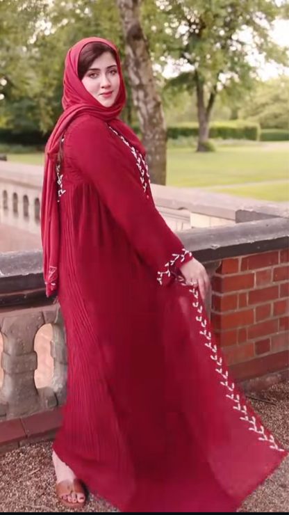 Woman in a red dress with white embroidery standing outdoors near a brick wall and trees.