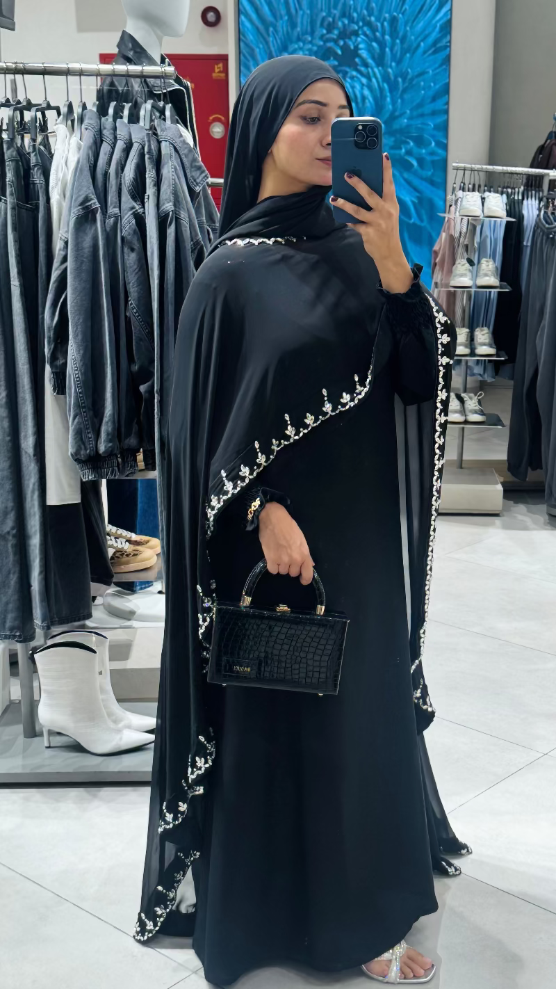 Woman taking a mirror selfie wearing a black abaya with white embroidery in a clothing store.