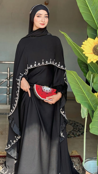 Woman in a black abaya with white embroidery holding a watermelon-themed clutch, standing next to a plant.