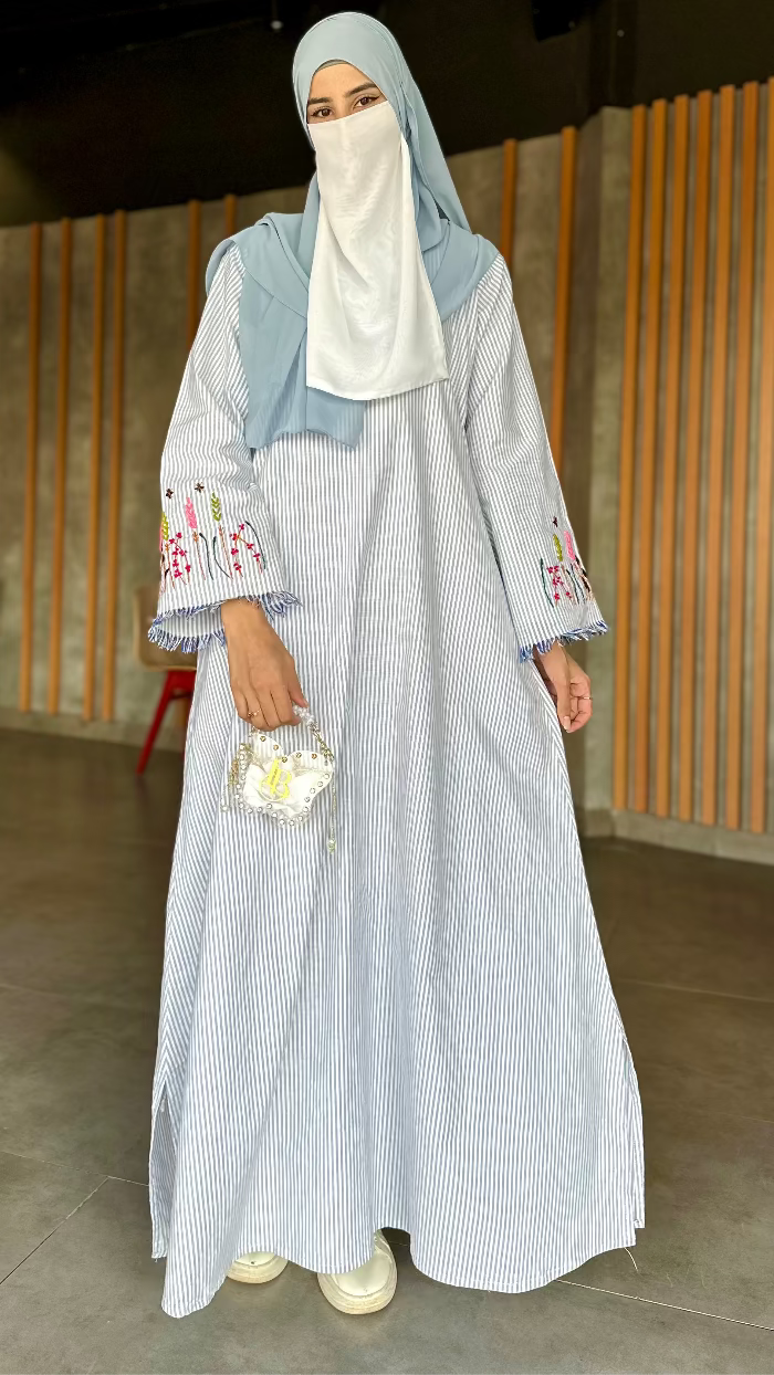 Woman in a light blue dress with floral embroidery standing indoors.