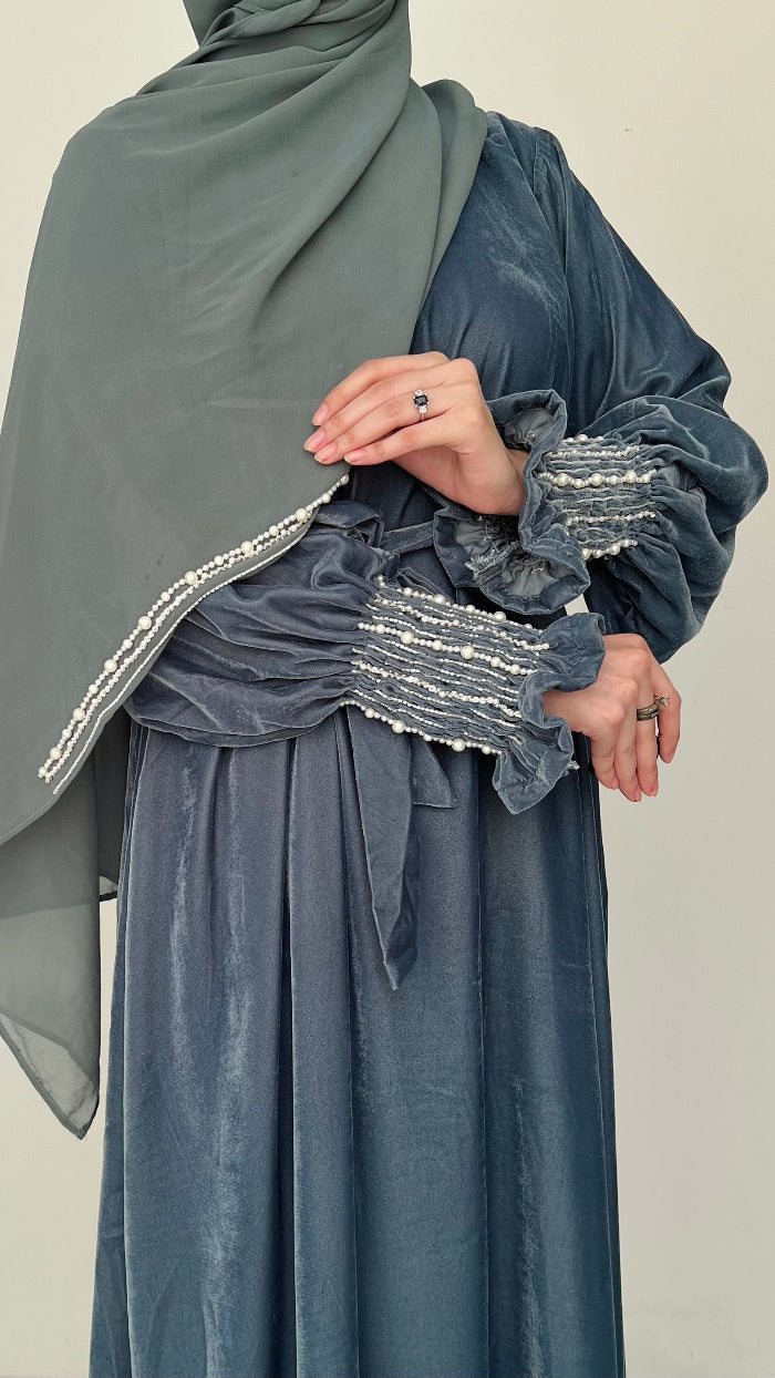 Person wearing a long, dark blue dress with ruffled sleeves and a matching hijab on a plain background