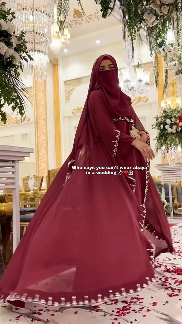 Woman in a red hijab and dress standing in a decorated indoor setting with flowers and lights.