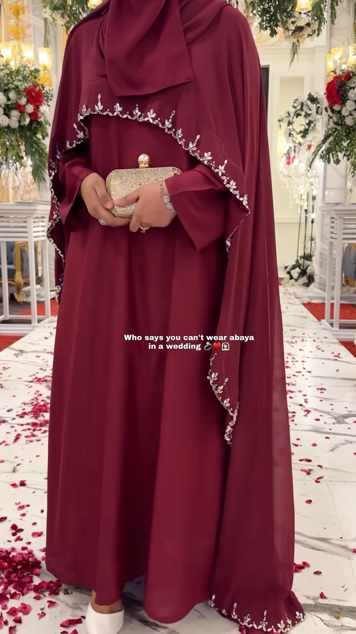 Person wearing a burgundy abaya with white embroidery in a decorated indoor setting.