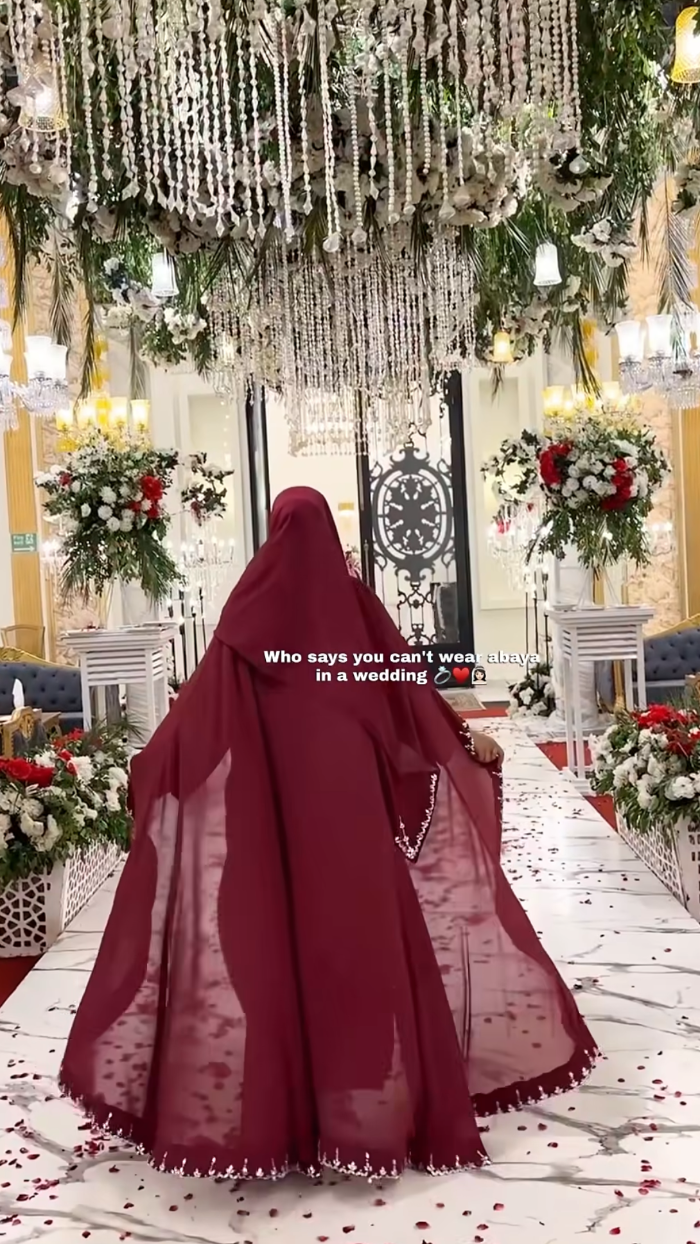 Person in a red hijab standing in a decorated room with chandeliers and floral arrangements.
