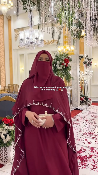 Woman in a red abaya standing in an ornate indoor setting with floral decorations.