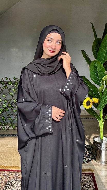 Woman wearing a dark gray abaya with pearl embellishments in an indoor setting with plants.