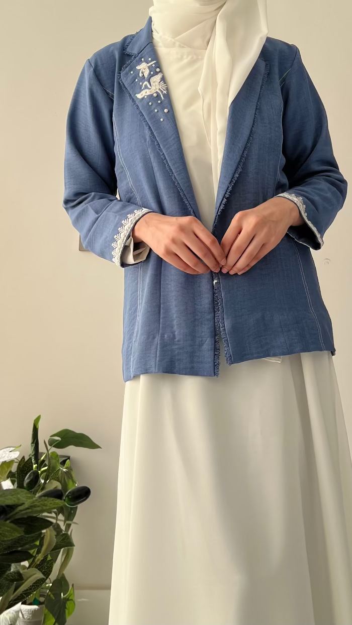 Person wearing a blue embroidered jacket with a white scarf and pants, standing next to a plant.