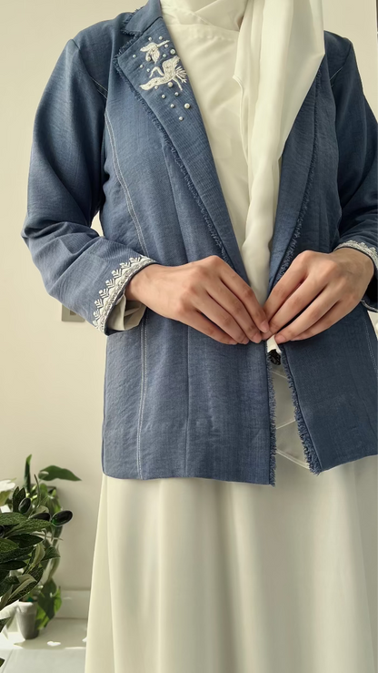 Person wearing a blue embroidered jacket with a white background and plant on the side