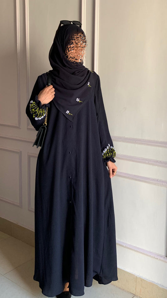 Person wearing a black abaya with floral patterns in a room with white paneled walls.
