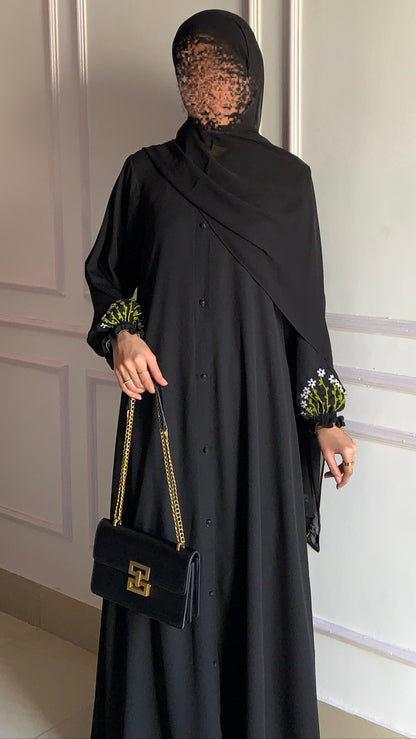 Woman in a black abaya with floral cuffs holding a black handbag against a white wall.