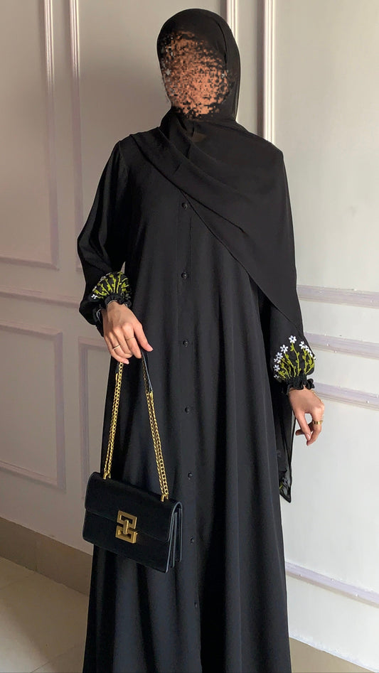 Woman in a black abaya with floral cuffs holding a black handbag against a white wall.