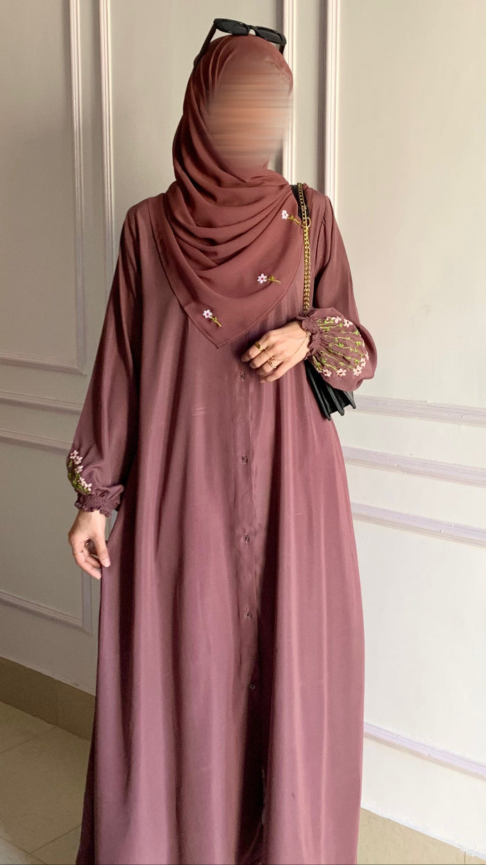 Person wearing a long mauve dress with a matching hijab against a white paneled wall.
