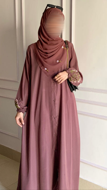 Person wearing a long mauve dress with a matching hijab against a white paneled wall.