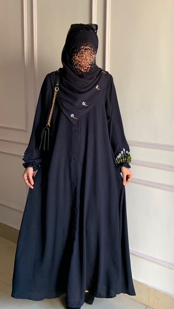 Person wearing a black abaya with floral embroidery in a room with white paneled walls.