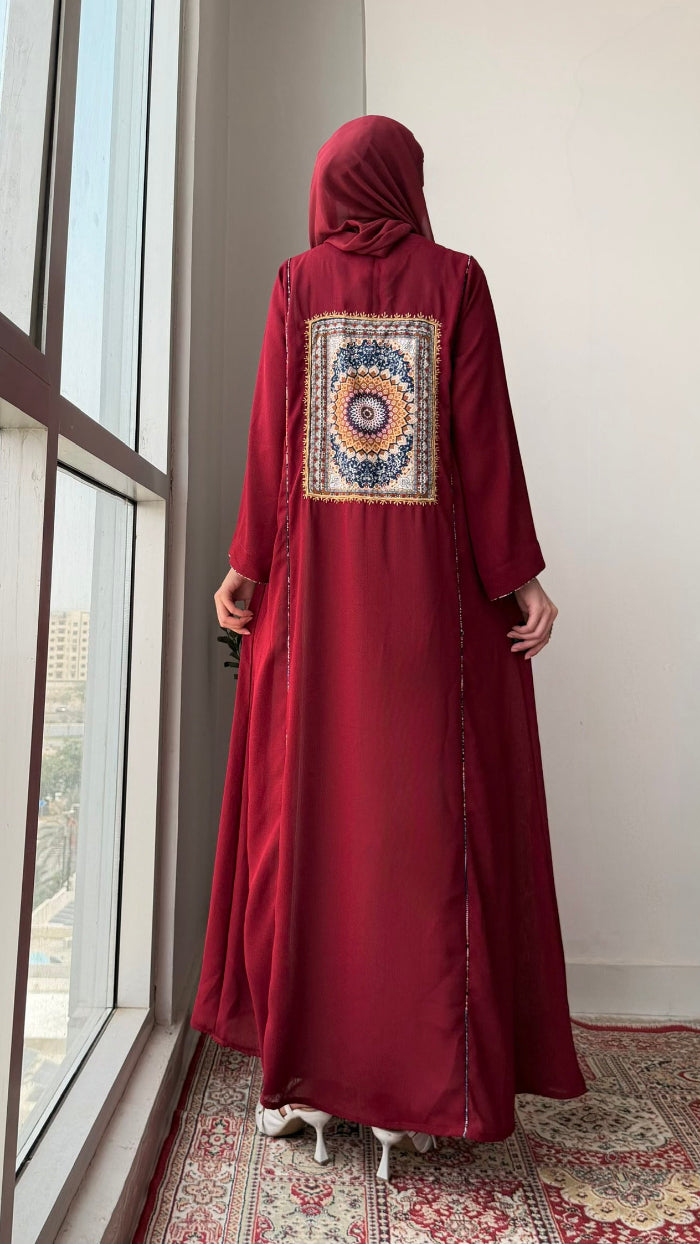 A woman stands with her back to the camera wearing a long maroon abaya with a large circular mandala pattern on the back panel, paired with a matching maroon hijab. She is standing on a patterned rug next to a large window.
