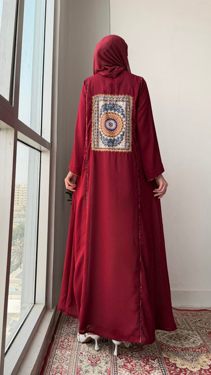 A woman stands with her back to the camera wearing a long maroon abaya with a large circular mandala pattern on the back panel, paired with a matching maroon hijab. She is standing on a patterned rug next to a large window.