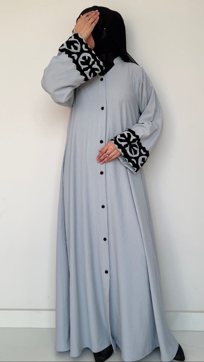 Woman wearing a long light gray dress with black buttons and cuffs, paired with a black hijab and gloves.