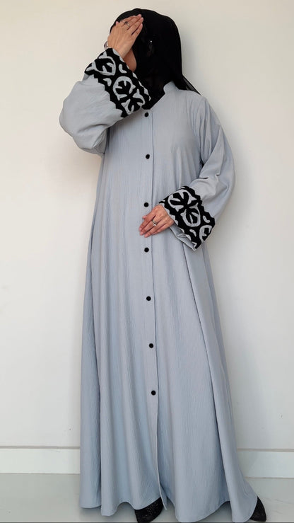 Woman wearing a long light gray dress with black buttons and cuffs, paired with a black hijab and gloves.