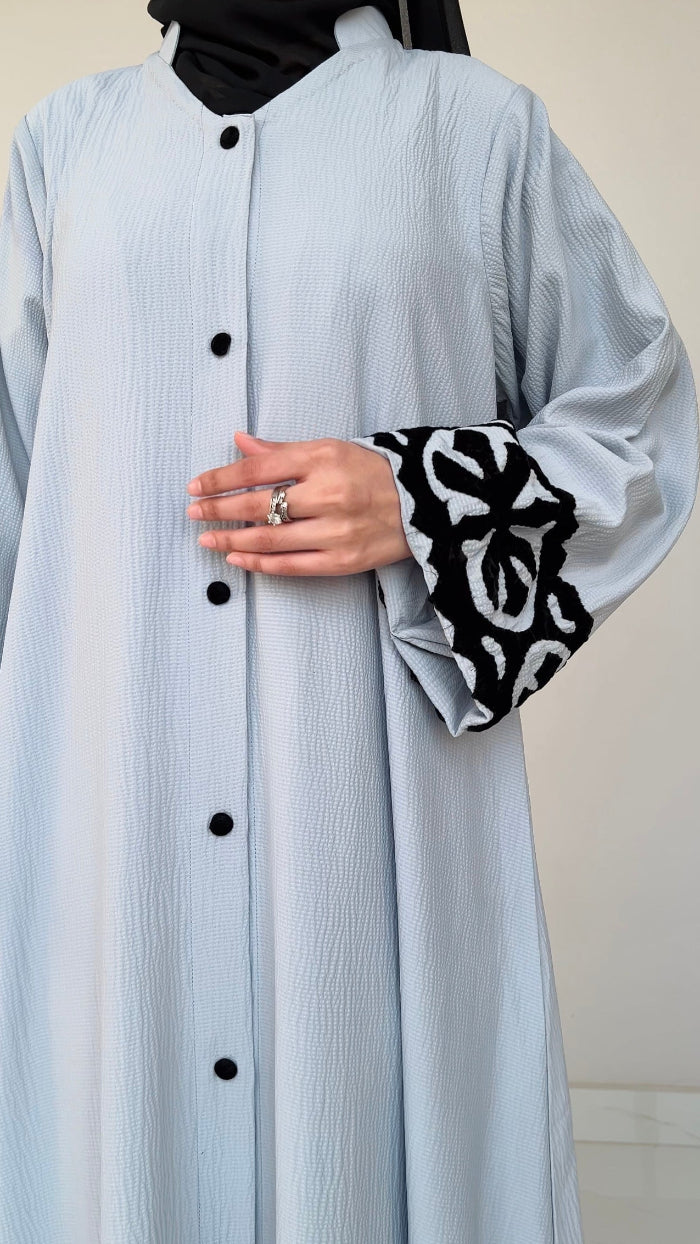 Light blue dress with black buttons and black and white patterned cuffs on sleeves against a neutral background