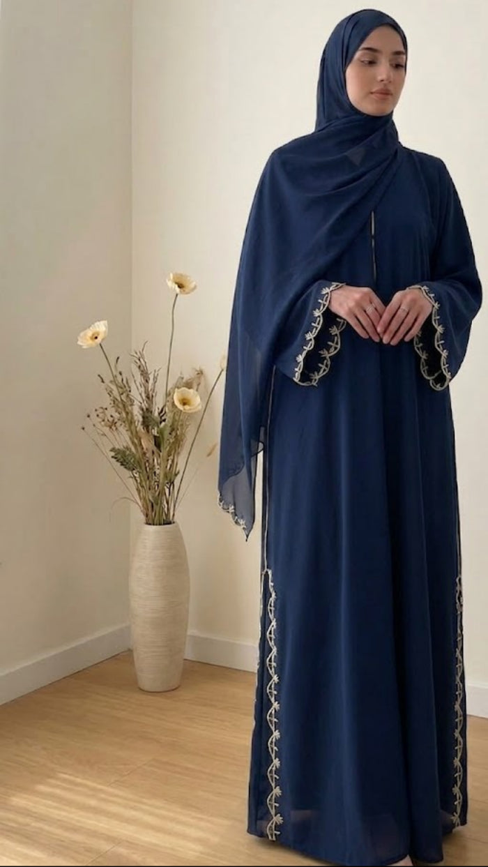 Woman wearing a dark blue abaya with intricate patterns standing in a room with a vase of flowers.