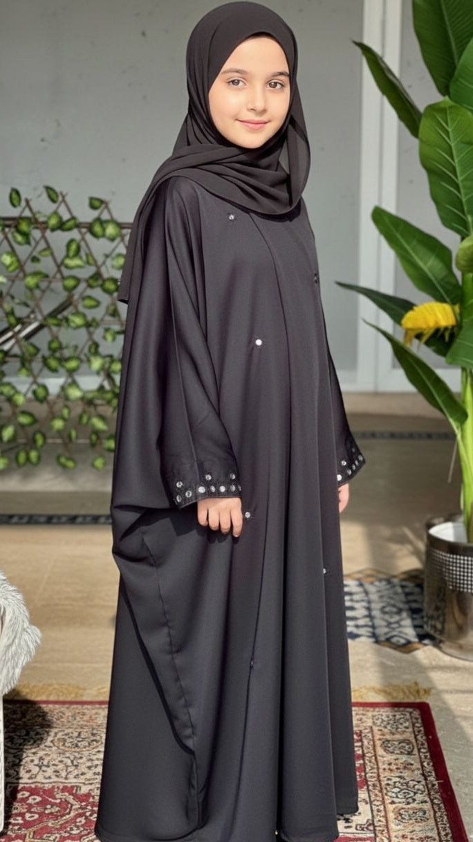 Young girl wearing a black abaya in a home setting with plants and decor.