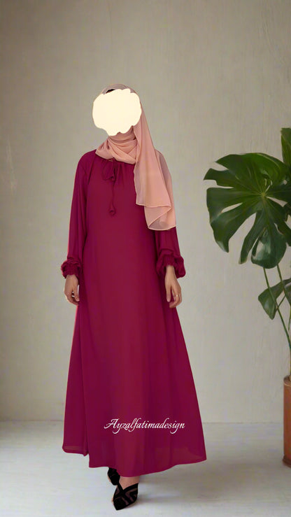 Person wearing a long burgundy dress with a beige hijab on a plain background